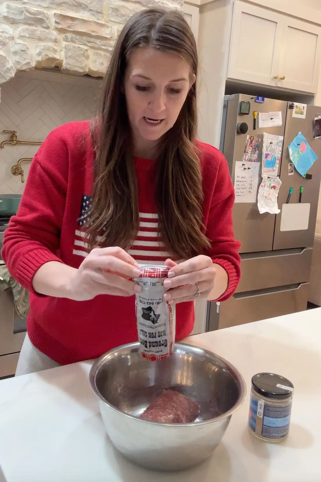 A woman in a red sweater with an American flag design is in a kitchen, opening a roll of ground beef over a mixing bowl for making carnivore burgers. The kitchen has a cozy, homey feel with a stone backsplash and fridge covered in children’s artwork.